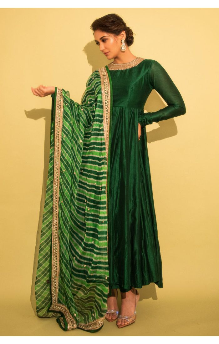 A model wearing a rich green Anarkali suit with a flowing silhouette, featuring full-length fitted sleeves and a satin-like finish. The outfit is paired with a green tie-dye patterned dupatta adorned with an embroidered border. Perfect for traditional cer